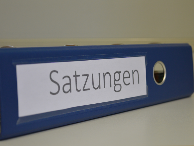 Ein blauer Ordner liegt auf einem weißen Tisch. Der Ordner ist mit der Aufschrift "Satzungen" beschriftet.