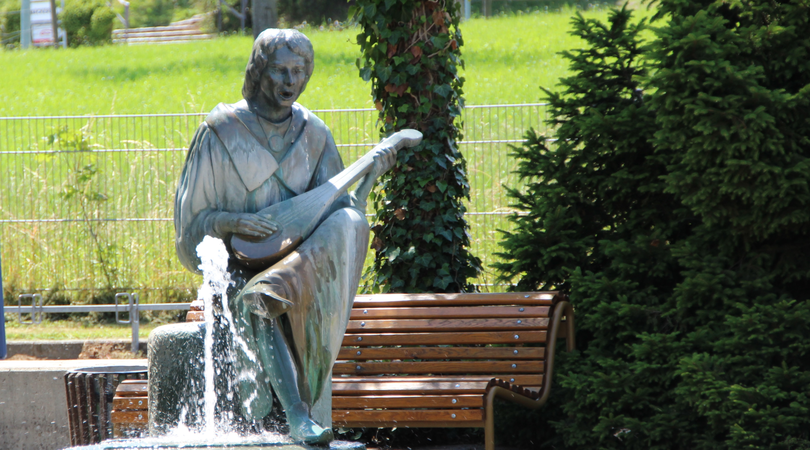 Ein Brunnen mit einer Wasserfontäne, der Brunnen hat die Form einer gitarrespielenden Frau